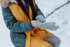 woman sitting outside in the snow wearing a blue coat, yellow scarf, and putting on gray gloves