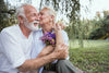 older couple sitting together in the grass, holding flowers and kissing