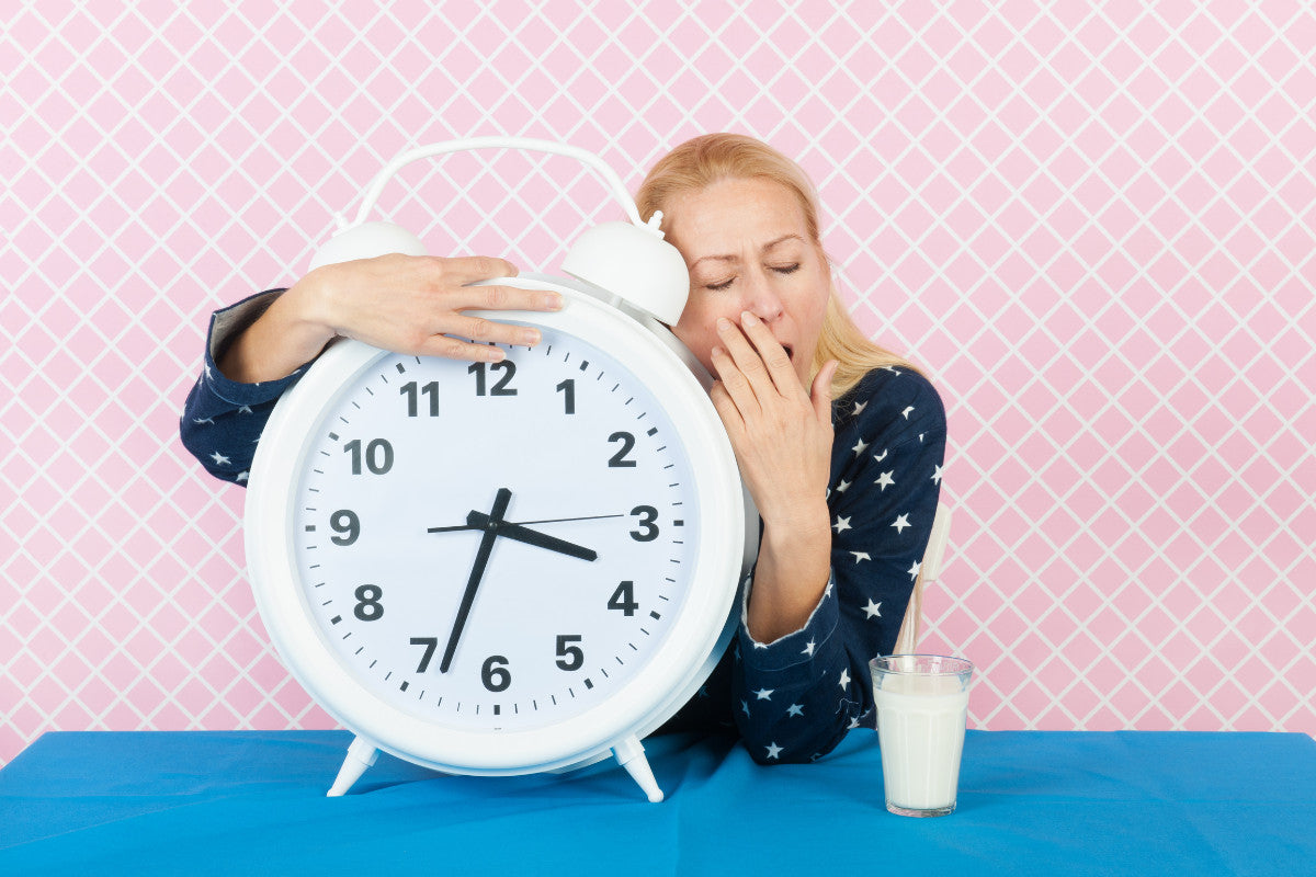 middle aged woman wearing pajamas, yawning, and holding a giant analog white clock.