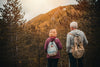 senior couple with their backs facing the camera, carrying small backpacks as they hike through a forest in the mountains during fall.