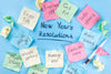 light blue background. pink, green, light blue, and pale yellow sticky notes with various New Year's Resolutions on them, surrounding the words 