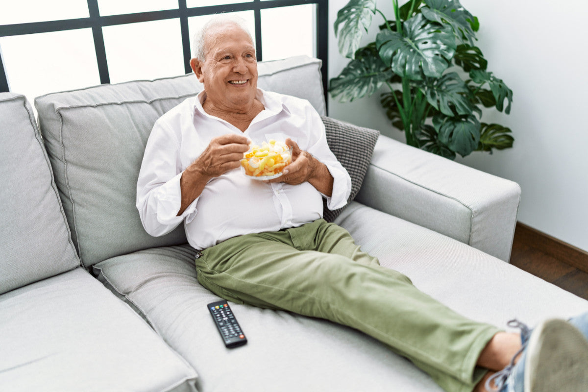 Senior man sitting on a gray couch, has his feet propped up as he eats chips out of a bowl.