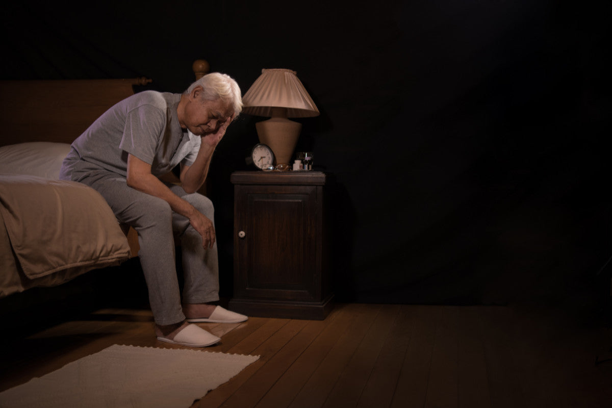 older man sitting on the edge of his bed wearing pajamas, struggling to sleep.