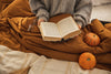 woman in gray sweater sitting under a brown comforter, reading a book with her glasses sitting next to her and two small orange pumpkins