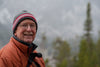 senior man smiling at the camera while standing outside during winter. standing in front of evergreen trees, wearing an orange coat, glasses, and a black beanie with a red and white stripe.