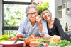 older couple acting silly while in the kitchen, various vegetables on the table in front of them