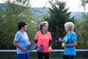three older women standing in a group on a tennis court, holding tennis rackets and tennis balls