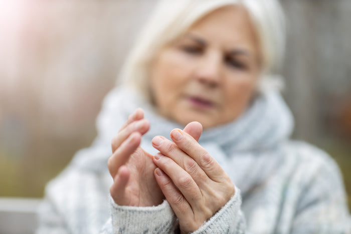 Senior woman with arthritis sitting outside on a park bench, wearing a winter coat, rubbing her hands together.