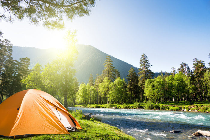 An orange tent set up next to a flowing stream in front of trees and mountains.