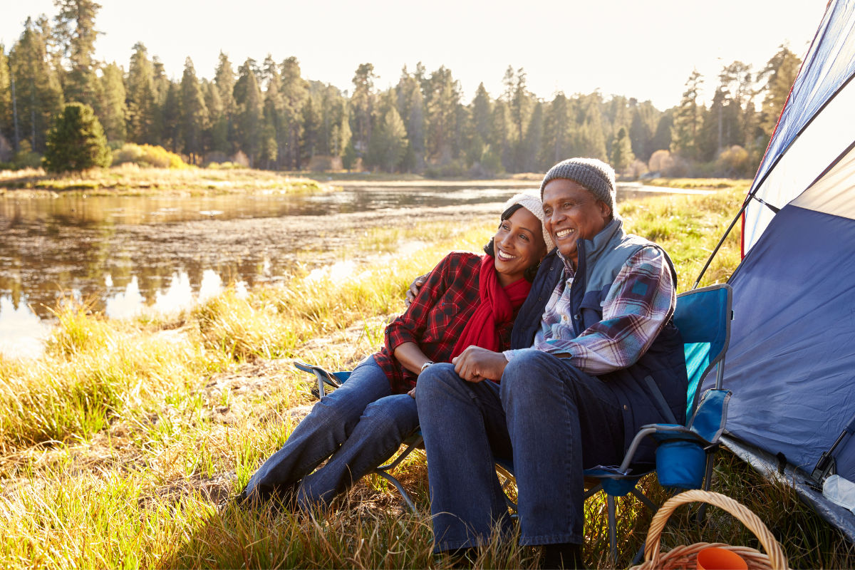senior couple sitting on grass next to a stream on lawn chairs and next to their white and blue tent