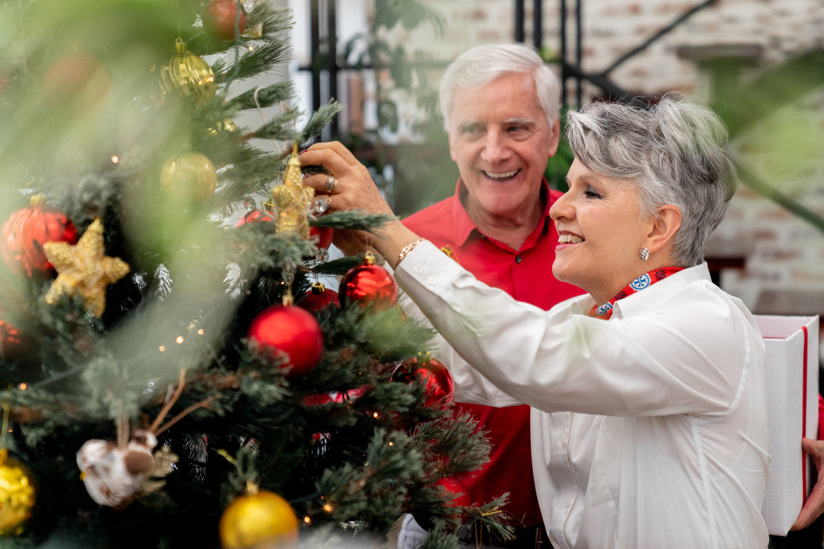 senior couple smiling as they decorate their Christmas tree with red and gold ornaments.