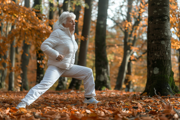 Older woman wearing a white coat, stretching outside in autumn leaves.