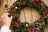 someone placing a small red and gold ornament on a wreath they are making. the wreath contains holly, pinecones, and red gold ornaments.