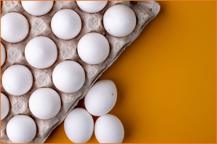 yellow background with a large gray container of white eggs with three white eggs sitting outside of the container