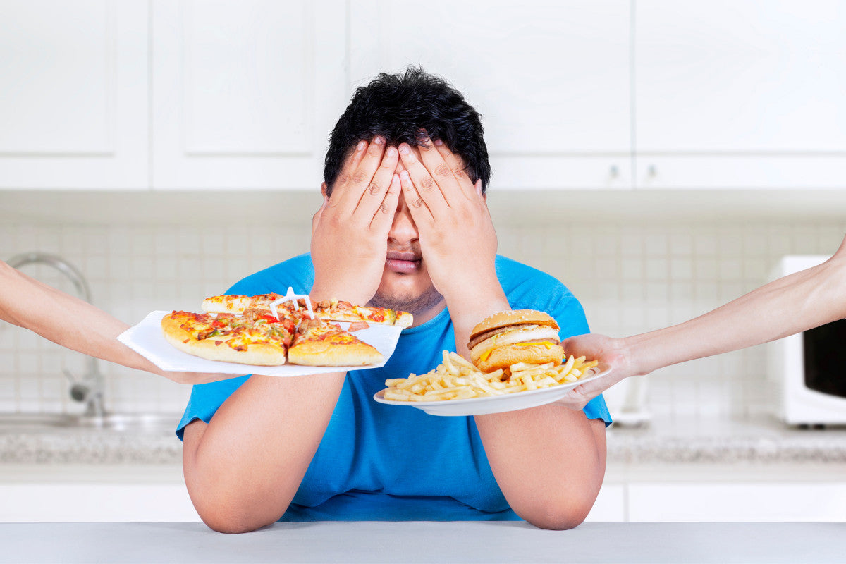 man wearing blue shirt, sitting at a white counter in a white kitchen, with his hands covering his eyes and forehead while two hands are holding out plates of junk food: one plate is pizza, and one plate is a hamburger and fries