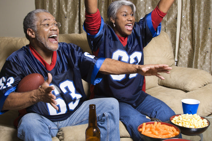 senior couple wearing football jerseys, cheering on the couch in their living room.