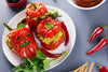 3 red peppers with jack-o-lantern faces carved into them, sitting on a white plate surrounded by red chili peppers and parsley.