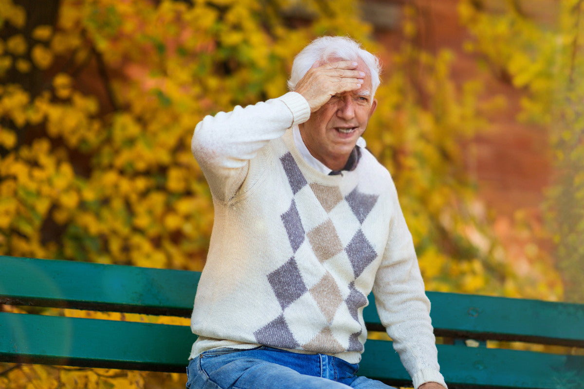 senior man sitting on a bench outside during autumn, his hand is covering his forehead as he suffers from a headache.