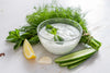 Cucumber and Mint Yogurt Dip in a glass bowl surrounded by mint leaves, cucumber slices, and a single lemon slice.