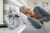 older woman laying in bed. alarm clock sitting on a white night stand in the foreground.