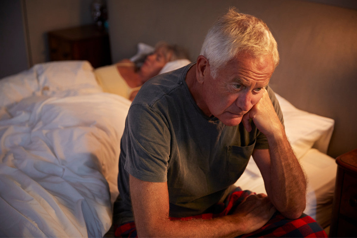 senior man sitting up in bed because he can't sleep while his wife sleeps peacefully next to him.