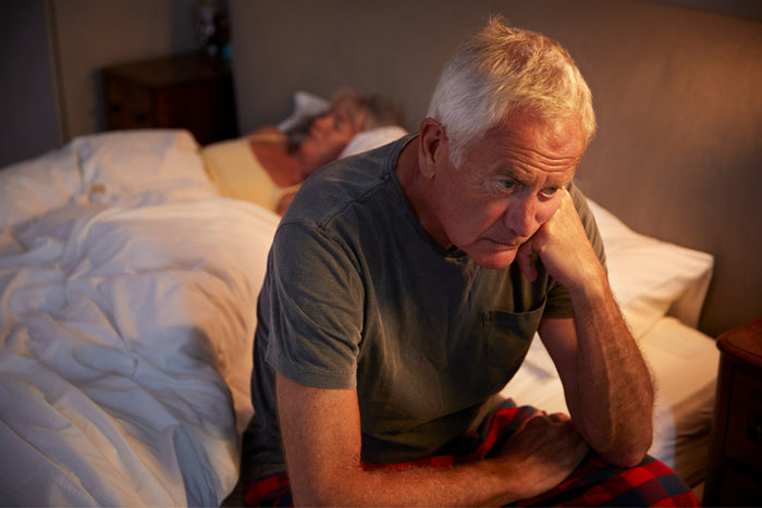 senior man sitting up in bed because he can't sleep while his wife sleeps peacefully next to him.