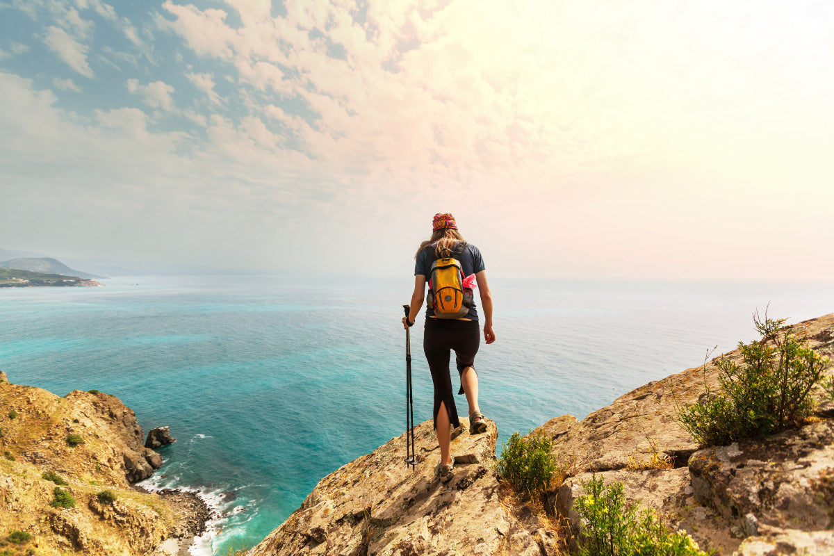 woman wearing hiking gear, standing a top a rock and looking out on the ocean.