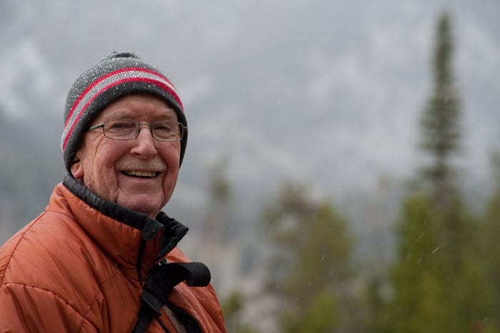 senior man smiling at the camera while standing outside during winter. standing in front of evergreen trees, wearing an orange coat, glasses, and a black beanie with a red and white stripe.