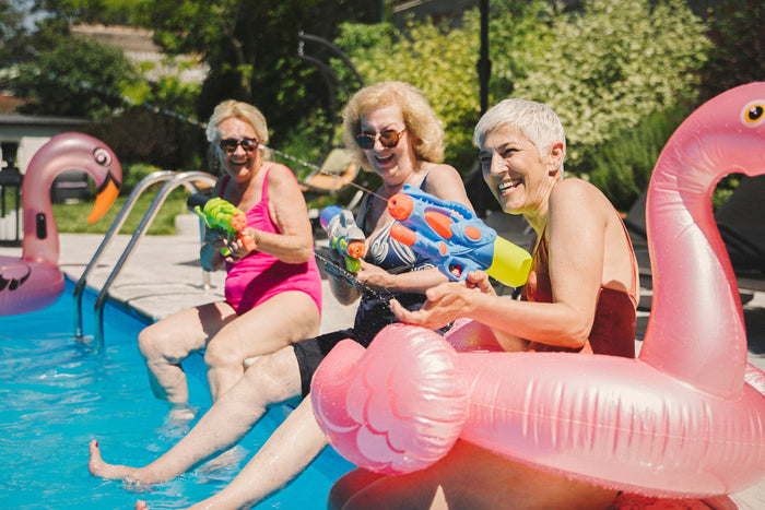 Mature women sitting on the edge of a pool spraying water guns at each other.