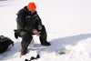 older man sitting on a white bucket in the snow with a fishing pole in his hand, wearing a dark green and black jacket and snow pants, and an orange beanie. ice fishing.