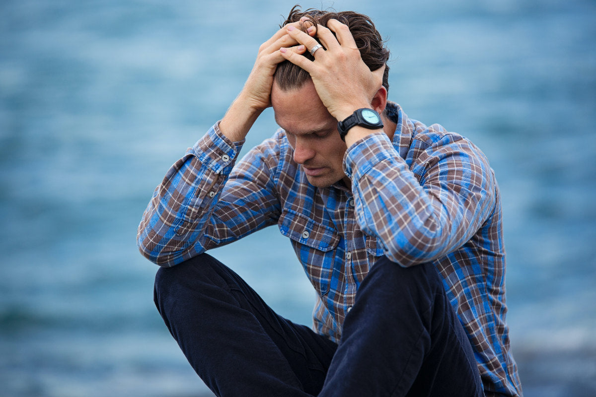 middle-aged man sitting cross-legged in front of a blue ocean background, with his hands on his head as if he's stressed.