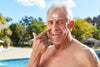 senior man smiling while applying sunscreen on his head and cheeks, standing in front of a pool during the summer